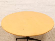Load image into Gallery viewer, A round light wood coffee table with a black metal swivel base, set against a white painted brick wall and light carpet.