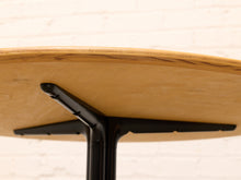 Load image into Gallery viewer, Close-up detail of a round table showing the light natural wood tabletop edge and black metal pedestal base against a white brick wall background.