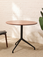 Load image into Gallery viewer, A round wooden table with a black metal tripod base, set on a textured beige rug against a white brick wall.