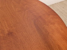Load image into Gallery viewer, Close-up of a round wooden table surface with a warm medium-brown finish and visible wood grain, with a small portion of a light rug and white baseboard visible in the background.