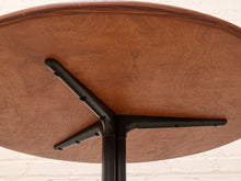 Load image into Gallery viewer, Close-up underside view of a round wooden table with a black metal pedestal base and three support legs, set against a white brick wall.
