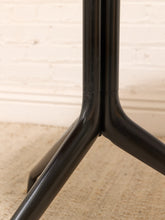 Load image into Gallery viewer, Close-up of a black metal tripod stand with three legs converging at a central joint, set against a light beige carpet and white brick wall background.