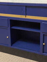Load image into Gallery viewer, A close-up of a blue cabinet or storage unit with a gold horizontal stripe, small gold knobs, and an open center shelf, shown against a neutral indoor background.