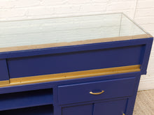 Load image into Gallery viewer, A close-up of a blue wooden display case or cabinet with a clear glass top, brass-toned metal trim, and a single drawer with a curved pull handle, set against a white brick wall.