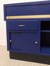 Load image into Gallery viewer, A close-up of a blue wooden cabinet or storage unit with a single drawer, two cabinet doors with round gold knobs, open shelving on the right, and a gold horizontal stripe near the top, set against a white brick wall and carpeted floor.