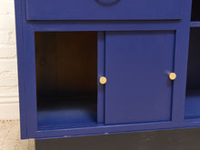 Load image into Gallery viewer, A close-up of a blue wooden storage cabinet or shelf with two small round gold knobs on the front door, set against a white brick wall background.