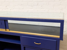 Load image into Gallery viewer, A blue wooden display case or cabinet with a glass top, gold trim, and a central gray panel, shown against a white brick wall.