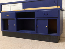 Load image into Gallery viewer, A navy blue wooden storage cabinet with a light wood top, featuring two drawers with brass handles, two lower cabinet doors with brass knobs, and an open center shelf, set against a white brick wall and beige carpet.