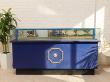 Load image into Gallery viewer, A modern blue display case or console table with a glass top, gold decorative trim, and a black base, set against a white brick wall with a potted plant nearby.