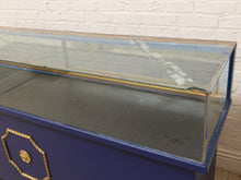 Load image into Gallery viewer, A vintage blue display case with a clear glass top and gold decorative trim, set against a white brick wall.