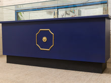 Load image into Gallery viewer, A custom blue display case or reception desk with a glass top, featuring a gold decorative octagonal frame on the front and a black base, set against a white brick wall.