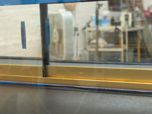 Load image into Gallery viewer, Close-up of a window or glass panel with a gold metal frame trim and blue painter's tape on the glass, with a blurred workshop or studio interior visible behind it.