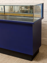 Load image into Gallery viewer, A blue fish tank display case with a clear glass top and a black base, shown against a white brick wall and beige carpet.
