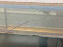 Load image into Gallery viewer, A close-up of a cracked clear glass surface, likely a tabletop or display case, with visible fracture lines and a dull metallic or bronze-toned edge along the bottom.