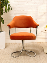 Load image into Gallery viewer, An orange velvet swivel dining chair with a chrome metal base, set against a white brick wall background with a potted plant on the left.