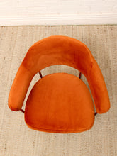 Load image into Gallery viewer, An orange velvet armchair with a curved backrest and seat, photographed from directly above on a beige woven rug against a white brick wall.