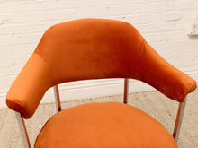 Load image into Gallery viewer, An orange velvet armchair with a curved backrest and slim metallic legs, positioned against a white brick wall and light beige carpet.