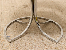 Load image into Gallery viewer, A close-up of a polished silver metal table base with a sculptural, curved leaf-like design, resting on a textured beige woven rug.