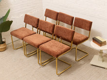 Load image into Gallery viewer, Six mid-century modern dining chairs with rust-brown corduroy upholstery and curved gold metal frames, arranged on a light beige carpet against a white brick wall.