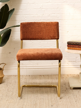 Load image into Gallery viewer, A mid-century modern barstool with a rust-orange corduroy upholstered seat and backrest, supported by a polished gold-finished metal frame, set against a white brick wall background.