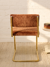 Load image into Gallery viewer, A modern cantilever chair with a rust-brown corduroy upholstered seat and back, supported by a polished gold metal frame, set against a white brick wall and light beige rug.