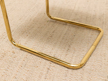 Load image into Gallery viewer, Close-up of a polished gold metal frame leg resting on a textured beige woven rug.