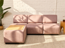 Load image into Gallery viewer, A blush pink modular sectional sofa with a matching ottoman in a bright, modern room with a white brick wall, natural light shadows, and a small wooden side table.