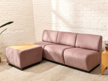 Load image into Gallery viewer, A modern L-shaped sectional sofa in dusty pink velvet with a matching ottoman, set in a room with a white brick wall and a light beige rug.