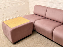 Load image into Gallery viewer, A mauve-pink modular sofa with a matching ottoman featuring a light wood-grain top, set against a white brick wall and beige textured rug.