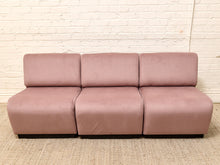 Load image into Gallery viewer, A three-seat modular sofa upholstered in dusty rose pink fabric, positioned against a white brick wall and a textured beige rug.