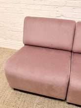 Load image into Gallery viewer, A dusty pink velvet modular sofa corner with a thick cushioned seat and backrest, set against a white brick wall and light beige woven rug.