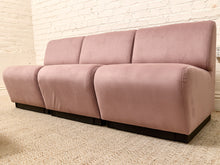 Load image into Gallery viewer, A three-seat sofa upholstered in dusty pink velvet with dark wooden legs, positioned on a textured beige carpet against a white brick wall.