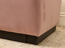 Load image into Gallery viewer, Close-up of the corner of a dusty pink upholstered ottoman or stool with dark wooden legs, sitting on a woven jute rug.