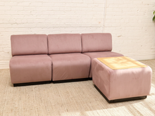 Load image into Gallery viewer, A three-seat modular sofa in dusty pink velvet with a matching ottoman featuring a light wood top, set against a white brick wall and beige rug.