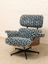 Load image into Gallery viewer, A mid-century modern lounge chair with a black, white, and blue geometric patterned fabric upholstery, a curved wooden shell, and a black five-star swivel base, set against a white brick wall and beige carpet.