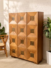Load image into Gallery viewer, A mid-century modern wooden cabinet with three doors, featuring a geometric, raised diamond pattern and square inset panels, set against a white brick wall with plants and a chair.