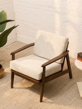 Load image into Gallery viewer, A mid-century modern accent chair with a dark brown wooden frame and light beige textured fabric cushions, set in a bright room with a white brick wall and a potted plant.