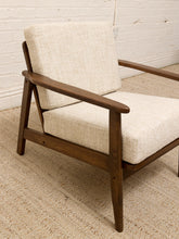 Load image into Gallery viewer, A mid-century modern wooden armchair with cream-colored textured fabric cushions, set on a woven jute rug against a white brick wall.