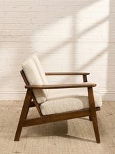 Load image into Gallery viewer, A mid-century modern accent chair with a warm brown wooden frame and textured cream bouclé upholstery, set against a white brick wall with soft window shadow patterns.