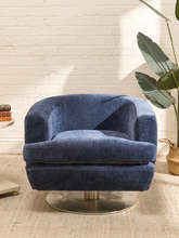 Load image into Gallery viewer, A blue velvet swivel armchair with a round metal pedestal base, set in a bright room with a white brick wall, a potted plant, and a woven rug.