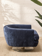 Load image into Gallery viewer, A modern barrel-style swivel armchair upholstered in deep navy blue velvet, mounted on a round brushed metal base, set against a white brick wall with a plant leaf in the foreground.