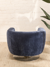 Load image into Gallery viewer, A modern barrel-style swivel chair upholstered in dark navy blue velvet, mounted on a round brushed metal base, set against a white brick wall and light beige rug.