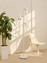 Load image into Gallery viewer, A modern white floor lamp with three adjustable cylindrical lights and gold-toned accents, standing in a bright room next to a white molded chair and a potted green plant against a white brick wall.