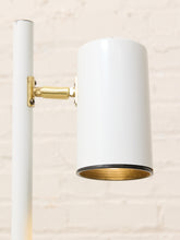 Load image into Gallery viewer, A modern wall-mounted floor lamp with a cylindrical white shade, a white vertical pole, and a polished gold metal bracket, shown against a light beige brick wall.