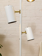 Load image into Gallery viewer, A modern white floor lamp with two adjustable cylindrical shades and brass-colored metal joints, set against a white brick wall background.