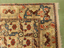 Load image into Gallery viewer, A close-up of a traditional patterned rug with a beige base, featuring red, blue, and brown floral motifs and a decorative border, laid on a green floor.