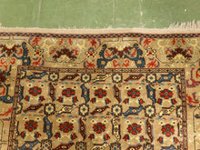 Load image into Gallery viewer, A close-up of a traditional patterned rug with a beige field, featuring red, blue, and brown floral motifs and a geometric border, laid on a green tiled floor.