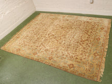 Load image into Gallery viewer, A rectangular vintage-style area rug with a faded beige and brown floral pattern and light blue border details, laid out on a green floor against a white brick wall.