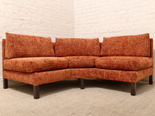 Load image into Gallery viewer, A curved three-seat sofa upholstered in textured rust-orange fabric, supported by dark wooden legs, set against a white brick wall and light beige carpet.