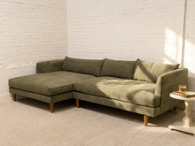 Load image into Gallery viewer, An olive green textured fabric sectional sofa with a left-facing chaise lounge and tapered wooden legs, set in a bright room with white brick walls and a light beige carpet.